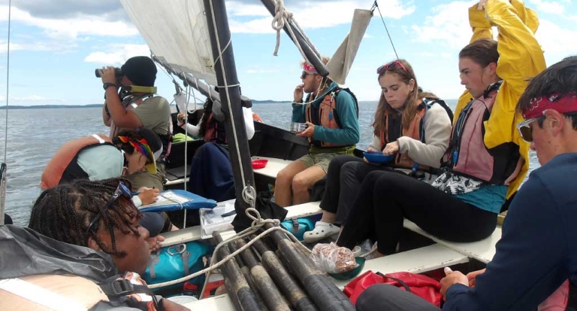 A group of students sit on a sailboat under blue skies. 